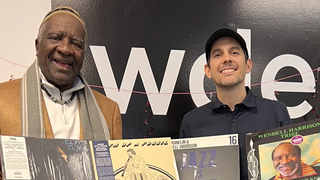 Wendell Harrison and Sam Beaubien smile and hold up some of Wendell's records in front of the WDET logo.