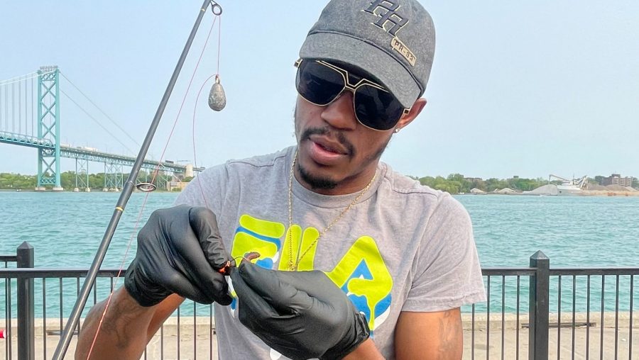 A man wearing a baseball hat, sunglasses and surgical gloves wraps a worm around his fishing hook
