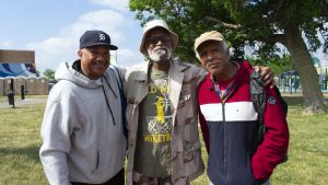 Three older Black men smile outside with their arms around each other