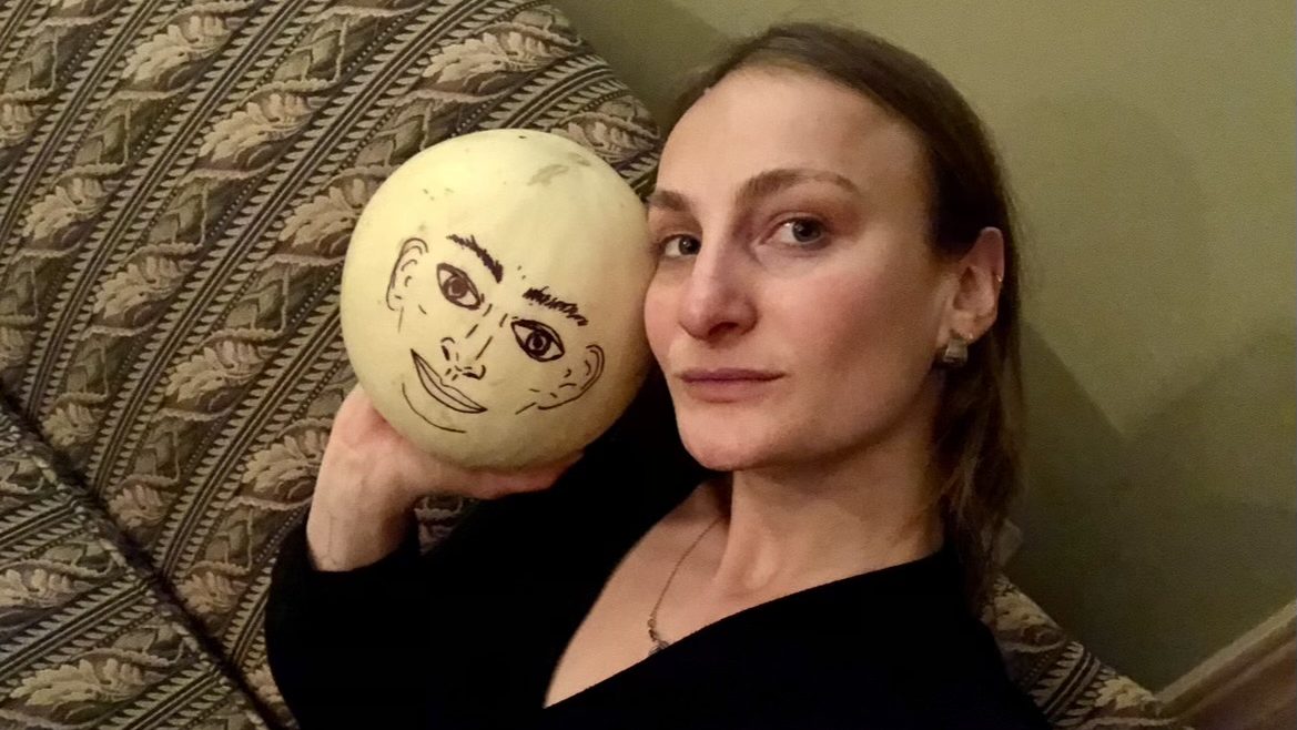 A woman poses on a couch holding a melon with a face drawn on it