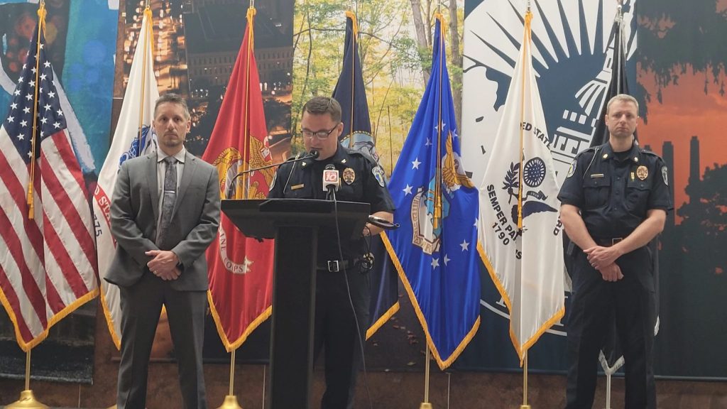 Lansing Police Chief Ellery Sosebee speaks into a micophone on a lectern, on his left is Supervisory Special Resident Agent Mark Civiletto with the FBI and on is right is Assistant Chief Robert Backus.