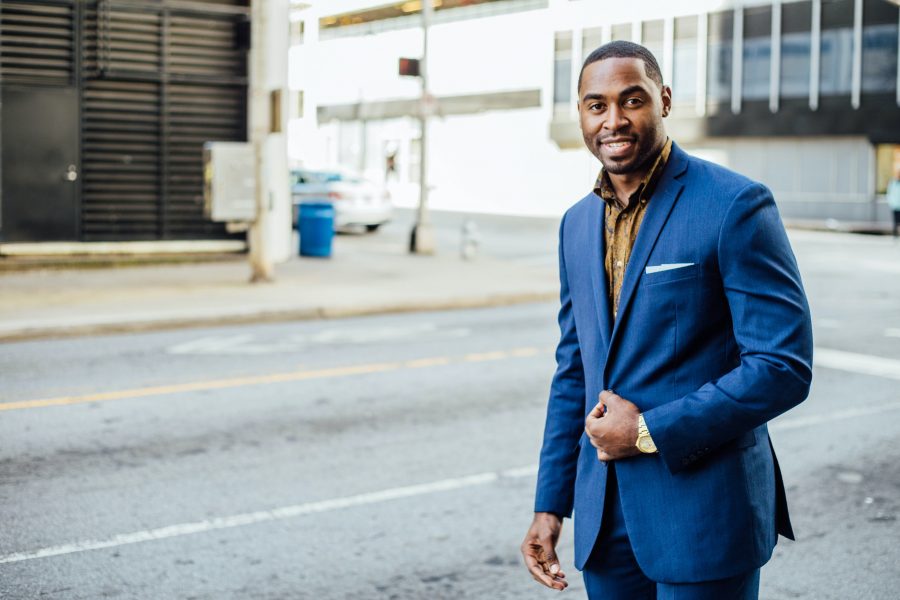 Photo of a Black man wearing a blue suit standing on the sidewalk.