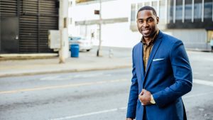 Photo of a Black man wearing a blue suit standing on the sidewalk.
