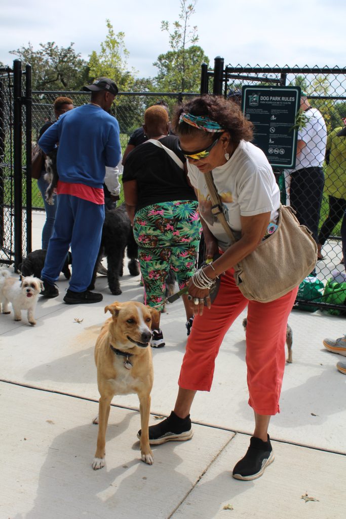 A dog owner wearing a headband, shades, and bag holds her dog on a leash in front of the gates to Palmer Park dog park.