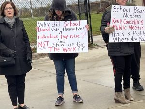 Teachers hold signs saying "Why did the school board vote themselves a raise and not show up for work?" and "School board members keep cancelling public meetings. That's a problem. where are you?"