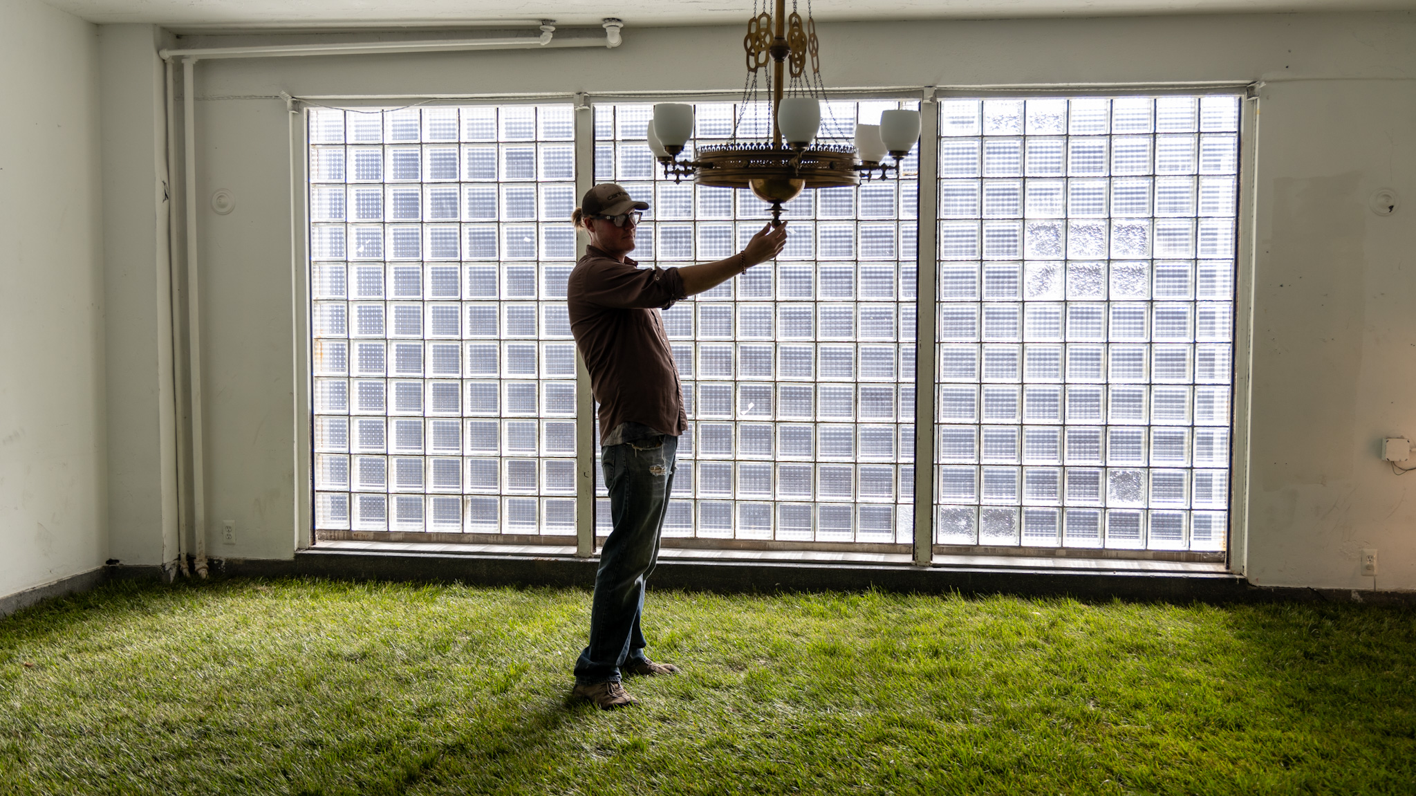A man lays looks at a light fixture while standing on the grass set inside the Boyer Campbell Building in Detroit for the Petrichor botanical art installation on May 30, 2024. (Photo credit: Tayler Simpson, WDET)