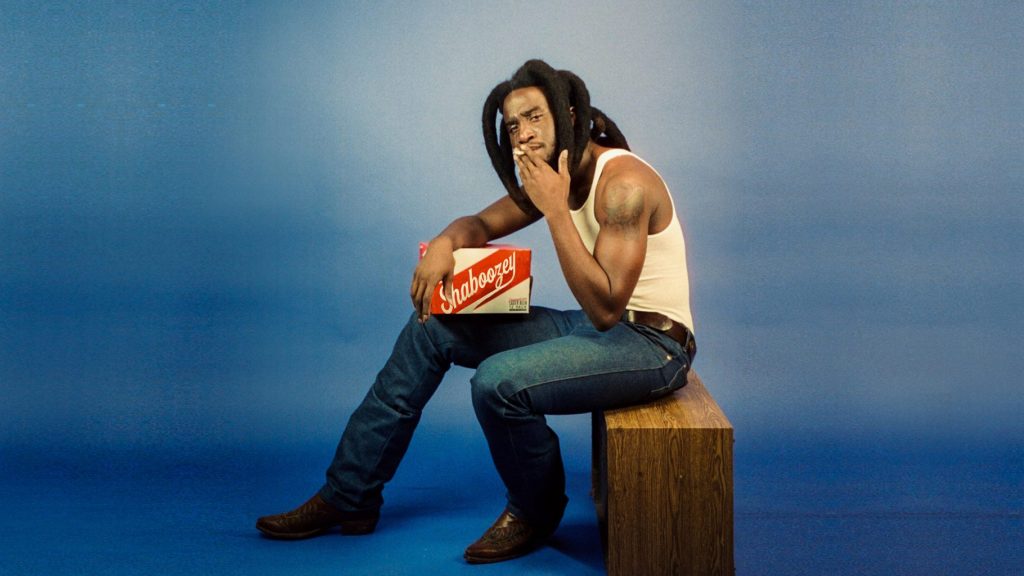 Shaboozey in blue jeans posing on bench.