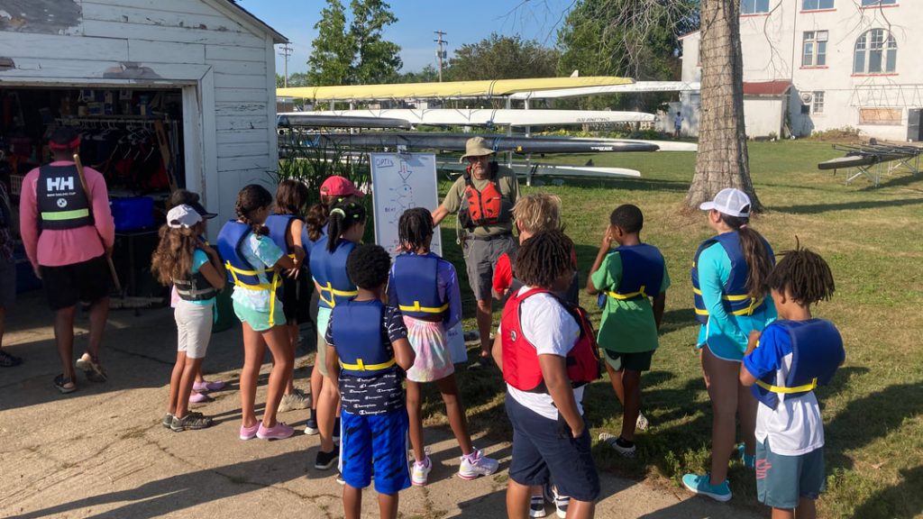 A group of kids in lifejackets stands around a white board for a sailing lesson