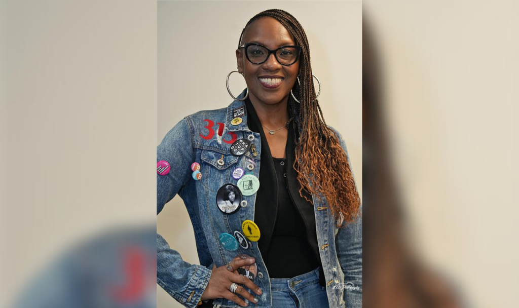 Kalimah Johnson stands confidently with a bright smile in a jean jacket laden with pins repping Detroit, social work, and other causes and organizations