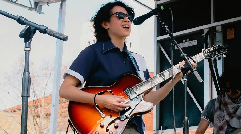 A woman in sunglasses plays an electric guitar with sunbrust paint job and sings at a microphone.