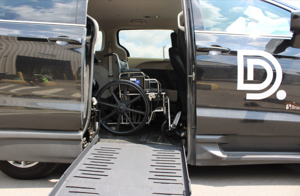 A mini van is parked with its side door open. A ramp is coming out the side and you can see a wheelchair in the van.