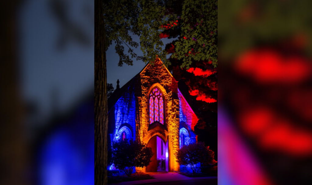 A stone building in a cemetery is lit up at night with purple, red and blue light.