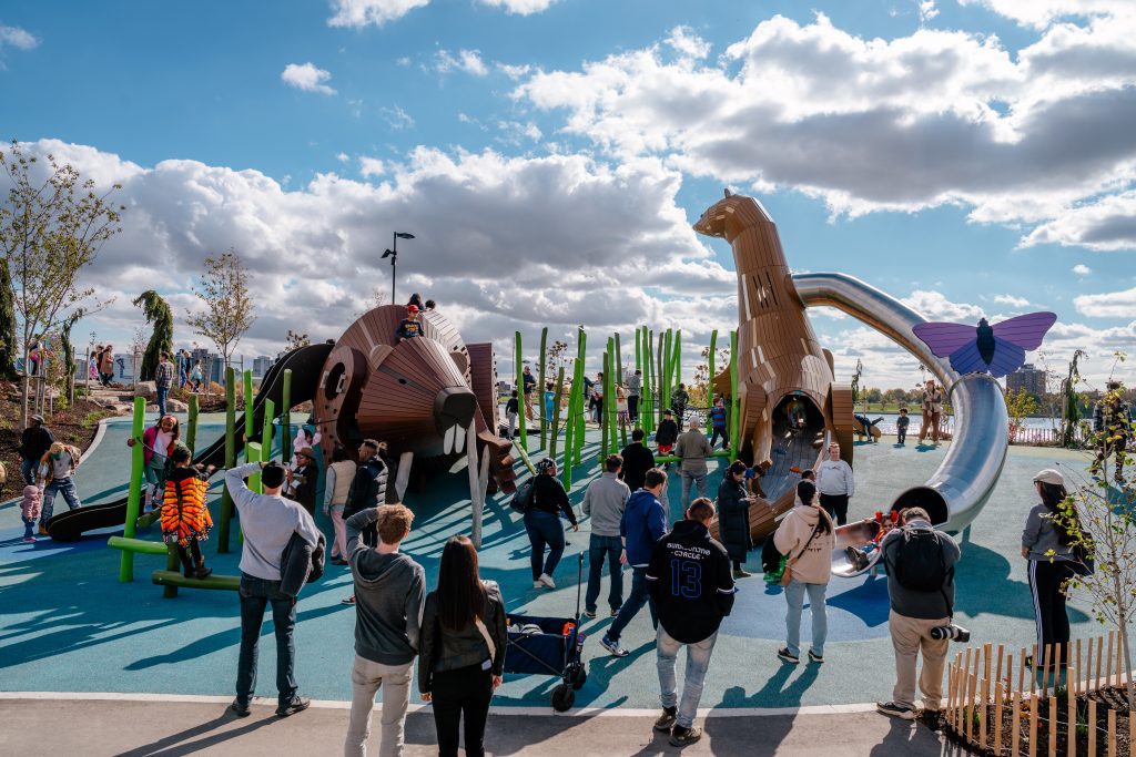Children and adults play and stand near playscape and splash pad at a public park in Detroit.