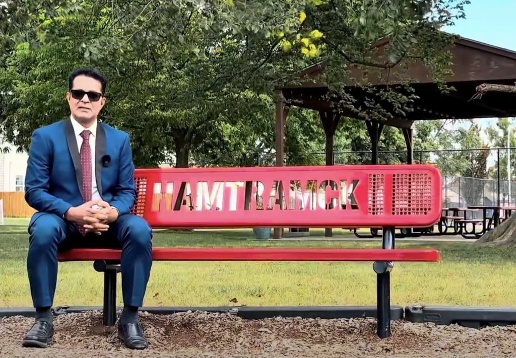 Hamtramck mayoral candidate Mahmood sits on a red bench that is designed to read "Hamtramck" on the backrest.