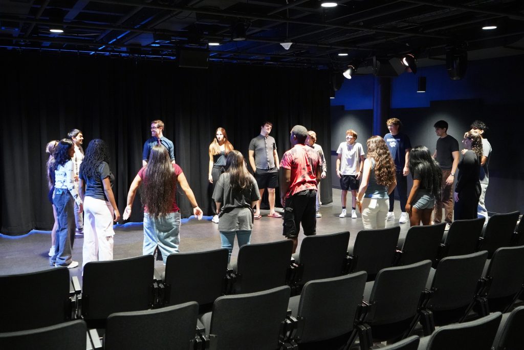 Actors stand in a circle in a dramatically lit theatre with black walls and cieling.