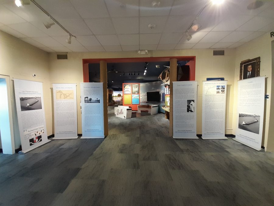Five pop up banners with photos and text of the Edmund Fitzgerald's Detroit story are on display in a museum with drop ceiling tiles and cream colored walls.
