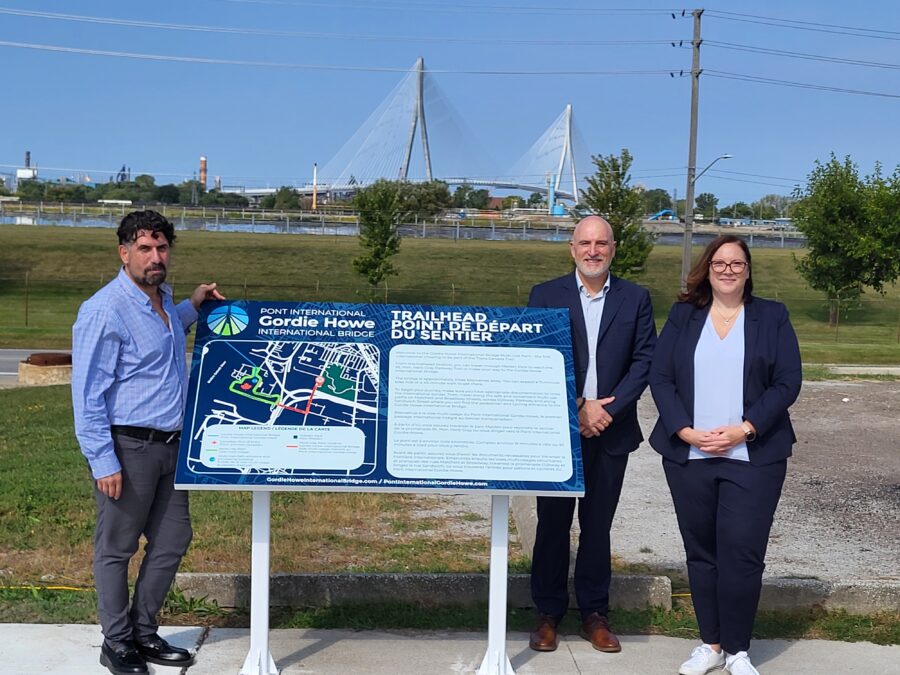 Three people pose next to a trailhead sign for the Gordie Howe International Bridge. The bridge is on the horizon in the background.